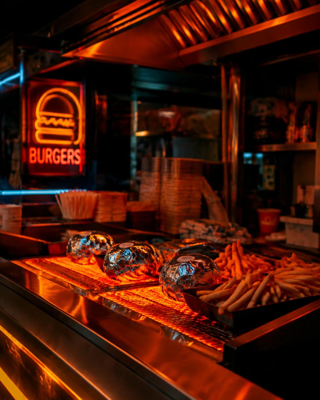 Neon-lit late night grill counter with foil-wrapped burgers and fries
